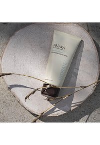 La mascarilla hidratante AHAVA en un tubo beige con tapa marrón, etiquetada como "HIDRATAR", descansa sobre una piedra circular clara con ramitas cerca.