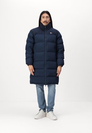 Námornícky puffer kabát s vysokým golierom, so zipsom vpredu, horizontálnym polstrovaním a logom na hrudi. Kombinovaný s svetlomodrými džínsami a bielymi teniskami.