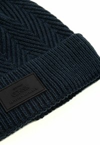 Ciemnoniebieska dziana czapka beanie z wzorami chevron i ściągaczami, z prostokątną czarną naszywką z wytłoczonym tekstem i współrzędnymi.