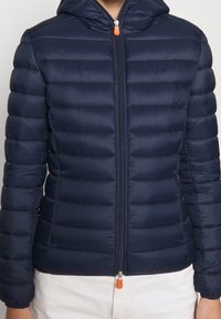 Marineblå pufferjakke med hætte, med quiltet struktur, frontlynlås med orange trækhåndtag og elastiske manchetter.