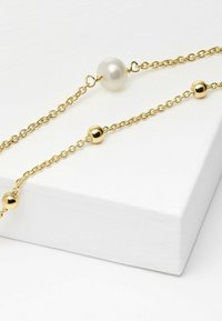 Collana in oro con una perla bianca centrale e accenti sferici in oro, posizionata su uno sfondo bianco texturizzato.