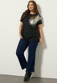 Sort t-shirt med et metallic guld sequin-design på den ene side, parret med mørkeblå flared jeans og ankelstøvler. Blødt stof, afslappet pasform.