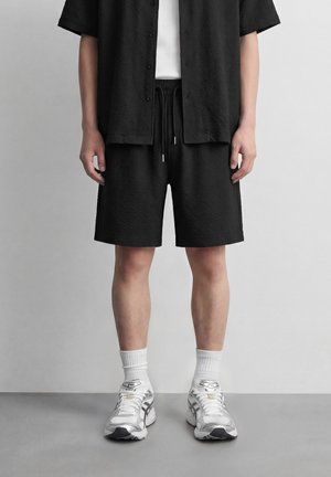 Personne portant un short texturé noir et une chemise à manches courtes assortie à boutons, des chaussettes blanches et des chaussures de course blanches, debout sur un sol gris.