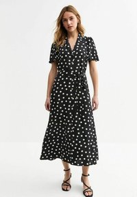 Vestido negro con lunares blancos, de mangas cortas, con botones en el frente, cinturilla anudada y longitud hasta la mitad de la pantorrilla. Presenta grandes lunares blancos y un diseño fluido.