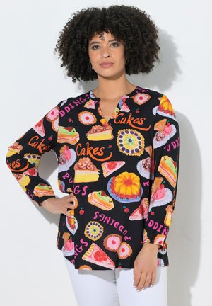 Woman with curly hair wearing a black long-sleeve shirt patterned with colorful cakes, pies, and the words "cakes" and "puddings."