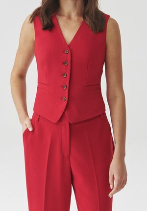 Vrouw draagt een mouwloos rood gilet met knopen en bijpassende hoge taillebroek, met één hand in de zak en een neutrale achtergrond.