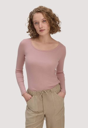 Junge Frau mit lockigem Haar, die ein hellrosa Langarmshirt und beige Hose trägt, mit den Händen in den Taschen, neutraler Hintergrund.