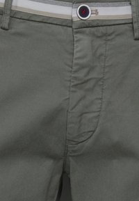 Pantalon vert olive coupe slim, avec texture sergé diagonale, poches latérales et fermeture avant à boutons avec un accent rouge.