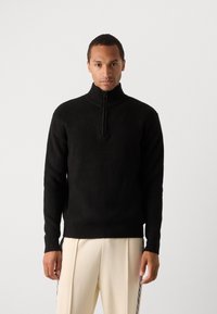 Svart strikket quarter-zip genser med høy krage og teksturert overflate, brukt sammen med kremfargede bukser med sidelister.