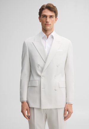 Uomo con capelli castano chiaro che indossa una giacca doppiopetto bianca e pantaloni abbinati, in piedi davanti a uno sfondo grigio semplice.
