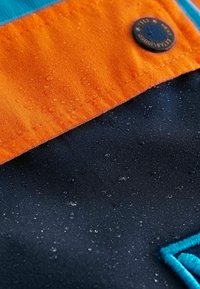 Vattentålig närbild av orange och marinblått tyg med synliga vattendroppar och en knapp på den orange sektionen.
