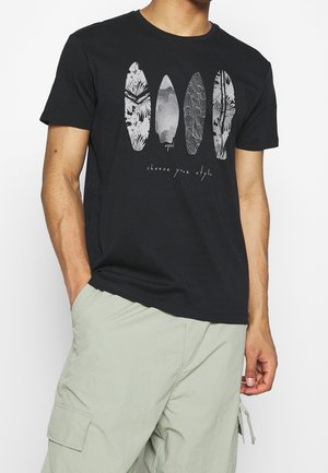 Homme portant un t-shirt noir avec quatre planches de surf stylisées et le texte « choisissez votre style », assorti à un short cargo vert clair.