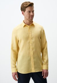 Camicia gialla a maniche lunghe realizzata in tessuto leggero, con colletto a camicia, bottoni frontali e orlo leggermente curvo.