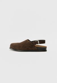 Καφέ παπούτσι από σουέτ με στρογγυλεμένη μύτη, χαμηλό τακούνι και ρυθμιζόμενο λουράκι με μεταλλική πόρπη στο πίσω μέρος.