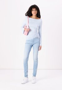 Top de manga larga azul claro con detalle fruncido, combinado con jeans skinny azul claro y zapatillas blancas. Llevando un bolso pequeño rosa.