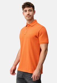 Narančasta polo majica s klasičnim ovratnikom, kratkim rukavima i suptilnim logotipom na prsima. Ima teksturiranu tkaninu i ravni rub.
