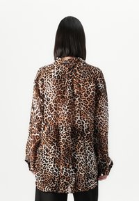 Pessoa com cabelo preto liso veste uma camisa de manga longa e folgada com padrão de leopardo, vista de costas contra um fundo branco simples.