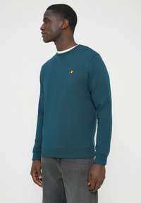 Tyrkysová mikina s dlhými rukávmi a okrúhlym výstrihom. Na hrudi žltá výšivka loga. Materiál z bavlnenej zmesi, voľný strih.