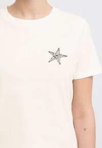 T-shirt en coton écru avec un graphique de starfis noir sur la poitrine gauche. Manches courtes et design classique à col rond.