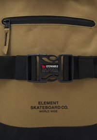 Brązowa torba z płótna z czarnym dnem, wyposażona w kieszeń na zamek, regulowany zamek z "Złożonymi paskami do deski skateboardowej z przodu" oraz tekstem logo.