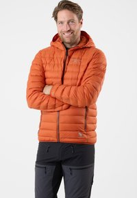 Dikke oranje geïsoleerde jas met een capuchon, een rits aan de voorkant en elastische manchetten. Draagt over donkere broeken met meerdere met rits afgesloten zakken.