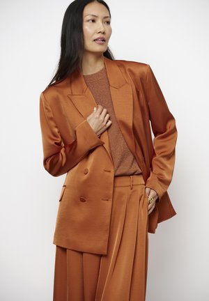 Vrouw met lang donker haar die een satijnen blazer in roestkleur draagt, bijpassende wijde broekspijpen en een gestructureerde top, poseert met één hand bij de kraag.