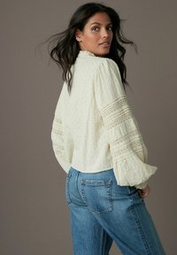 Blouse texturée couleur crème avec des manches volantes et des accents en dentelle, associée à un jean bleu délavé taille haute.