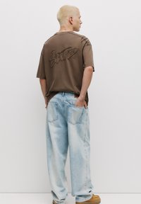 Hnědý oversized tričko s texturovaným designem na zádech, spárované se světle modrými volnými džíny a hnědými botami. Minimalistické pozadí.