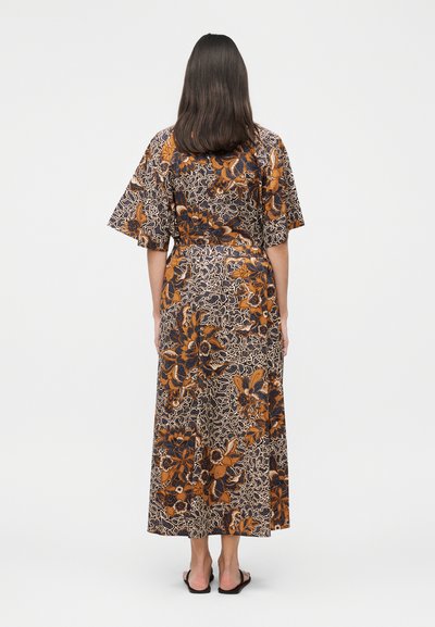 Γυναίκα με σκούρα μαλλιά έως τους ώμους που φορά ένα μακρύ καφέ και μαύρο φόρεμα με λουλούδια, στέκεται γυρισμένη προς τα πίσω σε λευκό φόντο.