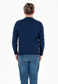 Granatowy sweter dzianinowy z teksturowanym kwadratowym wzorem, z prążkowanymi mankietami i dołem, w połączeniu z jasnoniebieskimi dżinsami. Widok z tyłu.