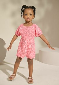 Rosa Floral-Overall mit kurzen puffärmel, gerüschtem Oberteil und gewelltem Saum. Kombiniert mit leichten Sandalen mit floralen Akzenten.