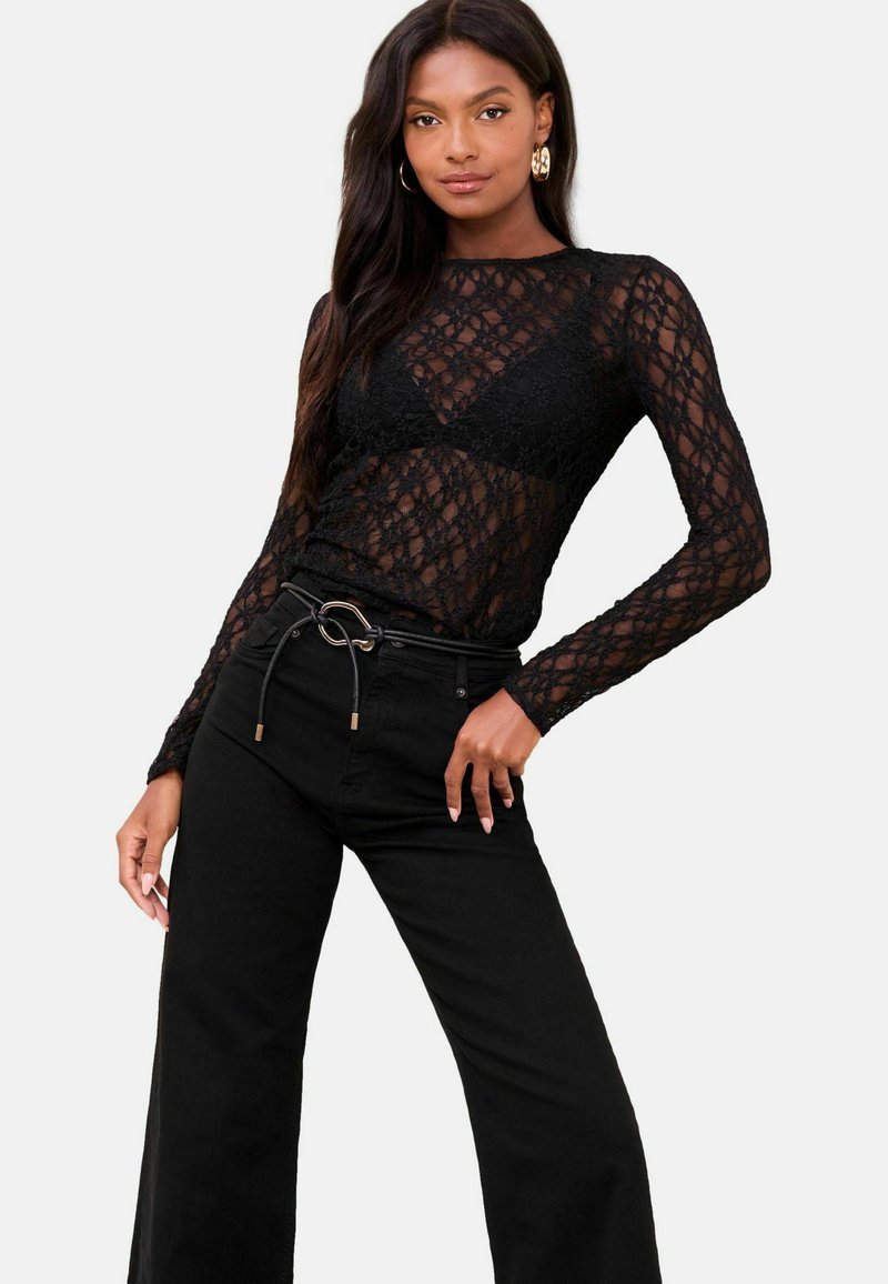 Kobieta ubrana w czarną koronkową bluzkę z długim rękawem na czarnej bralecie, w połączeniu z czarnymi spodniami z wysokim stanem i paskiem z okrągłą klamrą.