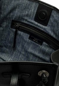 Intérieur d'un sac en cuir noir avec doublure en tissu denim sombre, une poche zippée et un bouton-pression rond noir.