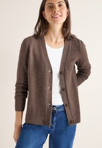 Brun, stickad cardigan med v-ringning, framknappar och ribbade manschetter. Bärs över en vit topp och tillsammans med blå denimjeans.