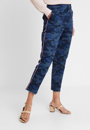 Vrouw draagt blauwe camouflage cropped broek met roze zijstrepen en beige sandalen met hak, hand in zak, staand op witte vloer.