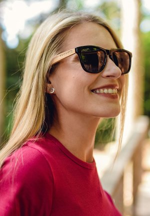 Black sunglasses with brown wooden arms featuring the engraved brand name "VUCH." Worn by a person with straight blonde hair, smiling.