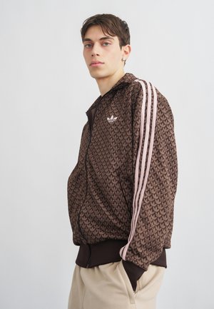 Νέος άνδρας που φοράει καφέ μπουφάν Adidas με ροζ ρίγες και μοτίβο τριφύλλι, ποζάροντας με τα χέρια στις τσέπες μπεζ παντελονιού μπροστά σε απλό φόντο.