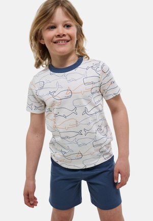 Enfant souriant portant un t-shirt blanc avec des dessins linéaires de baleines bleu marine et orange et un short bleu marine, debout devant un fond uni.