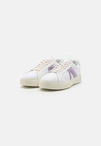 Zapatillas blancas con suela texturizada color crema, que presentan acentos en lavanda y gris en los laterales, y cordones color crema. Diseño limpio y moderno.