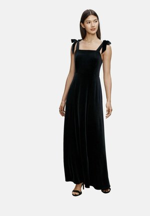 Schwarzes Samt-Maxikleid mit quadratischem Ausschnitt, gebundenen Schulterträgern und fließender Silhouette, kombiniert mit offenen schwarzen Absätzen.