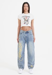 Cropped hvid t-shirt med grafisk print; lyseblå baggy jeans med gul tekst og logo detaljer; hvide sneakers; afslappet streetwear stil.