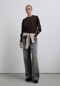 Femeie în picioare, îmbrăcată cu un top lung cu mânecă întunecată, blugi largi decolorati, sandale cu platformă și un pulover deschis legat în jurul taliei.