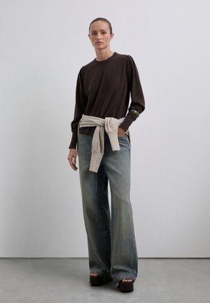 Frau steht in einem dunklen Langarmoberteil, weiten, ausgewaschenen Jeans, Plateausandalen und einem hellen Pullover, der um ihre Taille gebunden ist.