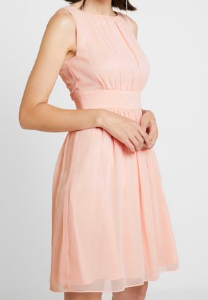 Kobieta w bezrękawowej, jasnoróżowej sukience do kolan z marszczonym talią i plisowanym dołem, stojąca na jednolitym tle.