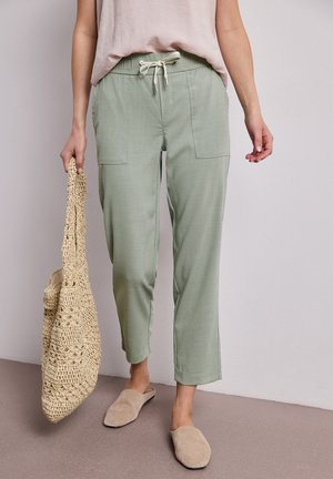 Kobieta ubrana w szałwiowe spodnie ze sznurkiem, beżowe buty wsuwane, trzyma plecioną beżową torbę na ramię, stoi na tle jednolitej ściany.