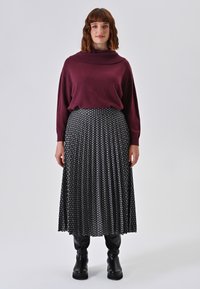 Bordeauxfarbener, oversized Strickpullover mit Kragen, kombiniert mit einem schwarzen Faltenrock mit weißem geometrischem Muster. Schwarze Knöchelstiefel.