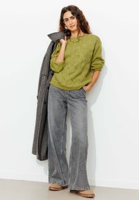 Pull en tricot vert avec un motif en vagues, associé à un jean gris à jambes larges et des bottes beiges. Un manteau à carreaux est posé sur un bras.