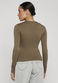 Donna con capelli scuri raccolti in uno chignon, indossa un top aderente a maniche lunghe color oliva e jeans grigio chiaro, vista di spalle su uno sfondo semplice.