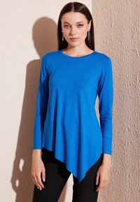 Top blu con maniche lunghe in tessuto leggero, con orlo asimmetrico, texture liscia e scollo rotondo, abbinato a pantaloni neri.