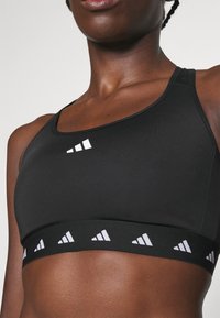 Sostén deportivo negro con un acabado texturizado, que presenta un logotipo blanco en el hombro y una banda con marca con un patrón de logotipo repetido en la parte inferior.